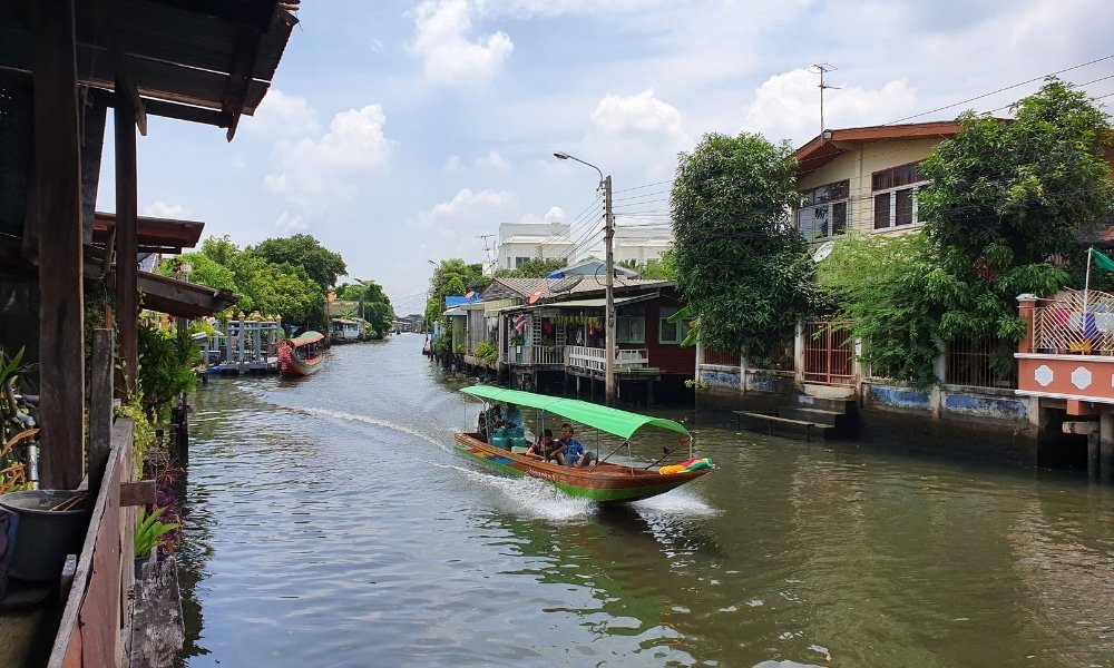 เรือหางยาวที่มีหลังคาสีเขียวแล่นไปตามคลองแคบที่เรียงรายไปด้วยบ้านเรือนและต้นไม้เขียวขจีใต้ท้องฟ้าที่มีเมฆบางส่วน มอบ ที่เที่ยวกรุงเทพ ทัศนียภาพของสถานที่ท่องเที่ยวในกรุงเทพมหานครสำหรับผู้ที่กำลังค้นหาความงามที่ซ่อนอยู่ของกรุงเทพฯ.