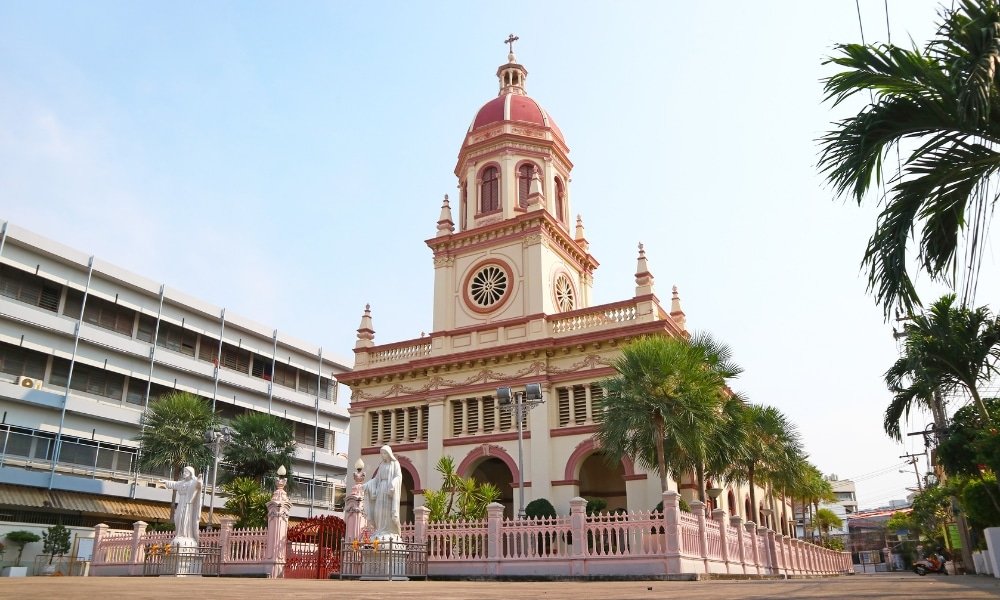 โบสถ์สีชมพูและครีมที่มีโดมใหญ่กลางและกางเขนตั้งอยู่ท่ามกลางต้นปาล์มและรูปปั้นภายใต้ท้องฟ้าที่มีเมฆบางส่วน ทำให้เป็นสถานที่ ที่เที่ยวกรุงเทพ ท่องเที่ยวที่ไม่เหมือนใครในกรุงเทพฯ สำหรับผู้ที่กำลังค้นหาสมบัติที่ซ่อนอยู่ของเมือง.