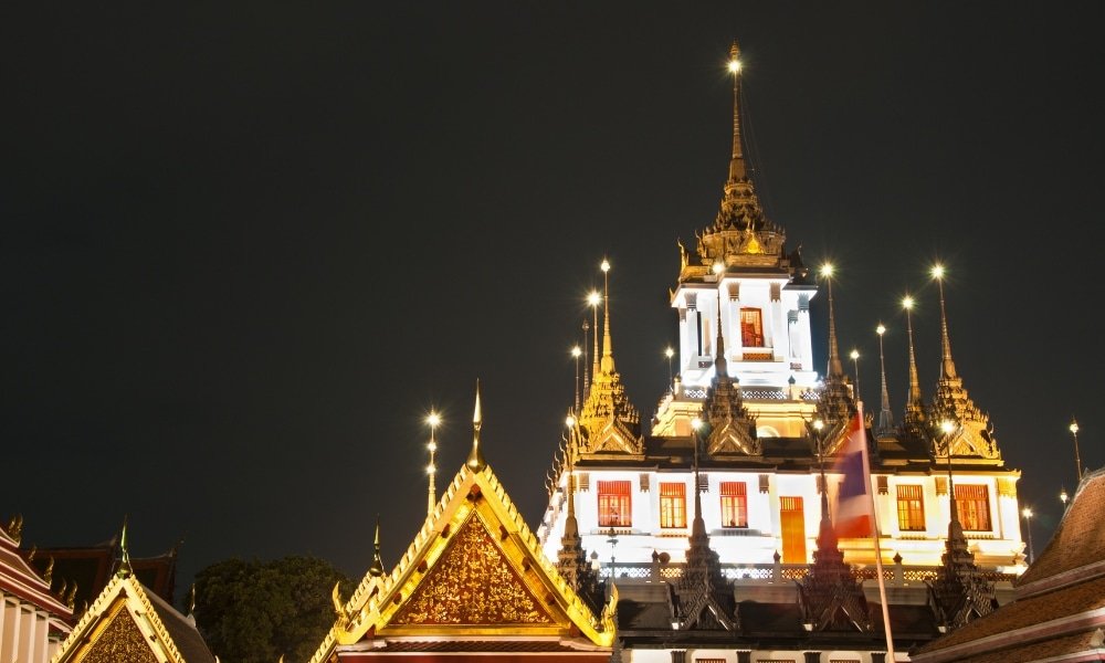 วัดไทยแบบดั้งเดิมที่ประดับไฟอย่างงดงามในยามค่ำคืน พร้อมยอดปราสาทที่ตกแต่งอย่างวิจิตร ที่เที่ยวกรุงเทพ ตระการตา ประดับด้วยสีทองและแสงไฟสวยงาม มีธงชาติไทยอยู่เบื้องหน้า—สถานที่ท่องเที่ยวกรุงเทพฯ ที่ไม่ควรพลาด.