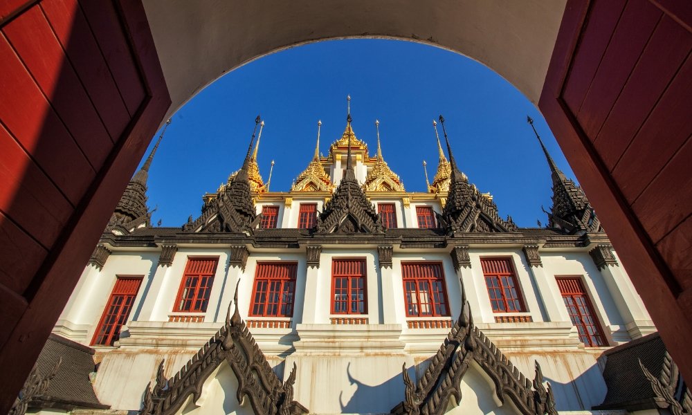 ภาพถ่ายแสดงให้เห็นวัดไทยที่ประดับประดาอย่างวิจิตร มียอด ที่เที่ยวกรุงเทพ เจดีย์สีทองและกรอบหน้าต่างสีแดง มองผ่านประตูโค้งที่มีท้องฟ้าสีฟ้าใสเป็นฉากหลัง—เป็นตัวอย่างอันงดงามของสถานที่ท่องเที่ยวในกรุงเทพฯ สำหรับผู้ที่กำลังสำรวจมรดกทางวัฒนธรรมอันล้ำค่าของเมือง.