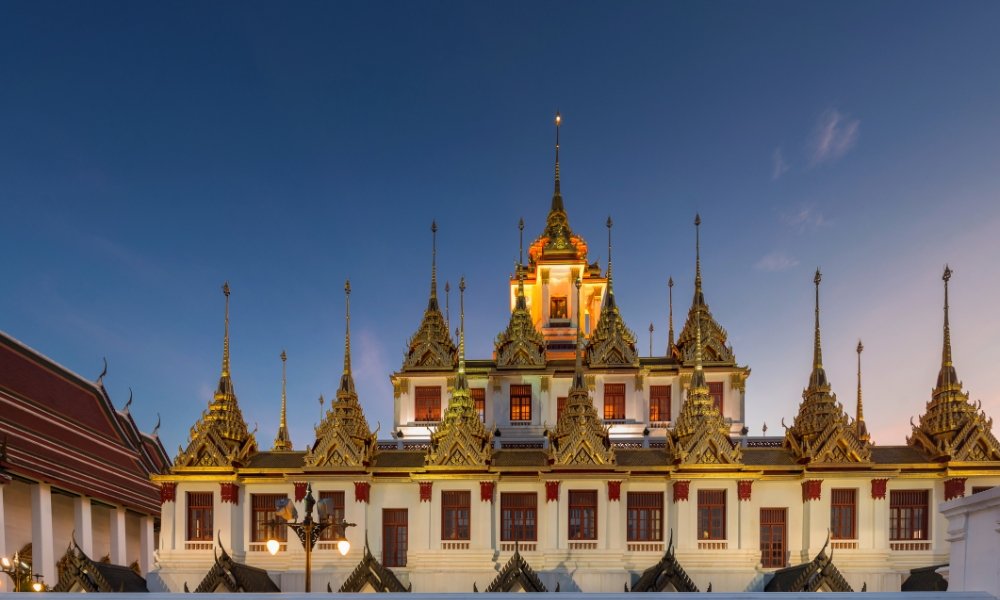 อาคารวัดที่ประดับประดาอย่างวิจิตรด้วยยอดแหลมสีทองและราย ที่เที่ยวกรุงเทพ ละเอียดที่ซับซ้อน ส่องแสงสว่างในยามพลบค่ำตัดกับท้องฟ้ายามค่ำคืนที่แจ่มใส—สถานที่ท่องเที่ยวในกรุงเทพฯ ที่ไม่ควรพลาด.