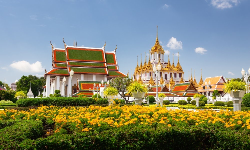 อาคารวัดไทยแบบดั้งเดิมที่มีหลังคาประดับประดาอย่างวิจิตรตั้งอยู่ท่ามกลางสวนที่ตกแต่งอย่าง ที่เที่ยวกรุงเทพ ประณีตและดอกไม้สีเหลืองภายใต้ท้องฟ้าสีคราม—สถานที่ท่องเที่ยวที่สงบในกรุงเทพฯ สำหรับผู้ที่ต้องการสำรวจมรดกทางวัฒนธรรมของเมือง.