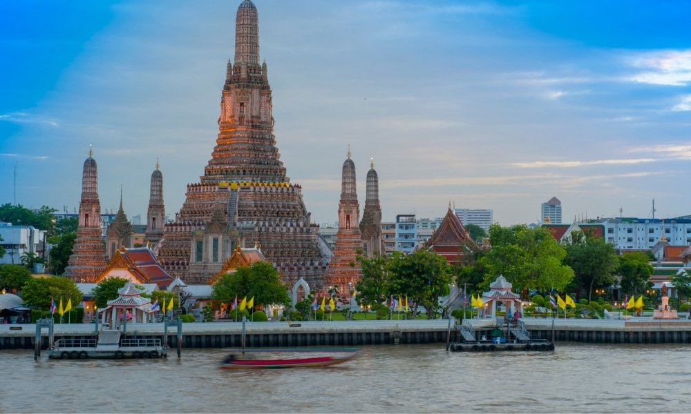 วัดอรุณราชวราราม วัดที่มียอดสูงตระหง่าน ตั้งอยู่ริมฝั่งแม่น้ำเจ้าพระยาในกรุงเทพมหานคร  ที่เที่ยวกรุงเทพ ประเทศไทย ทำให้เป็นสถานที่ท่องเที่ยวที่ต้องไปเยือนในกรุงเทพ พร้อมต้นไม้และตึกในฉากหลังภายใต้ท้องฟ้าสีคราม.