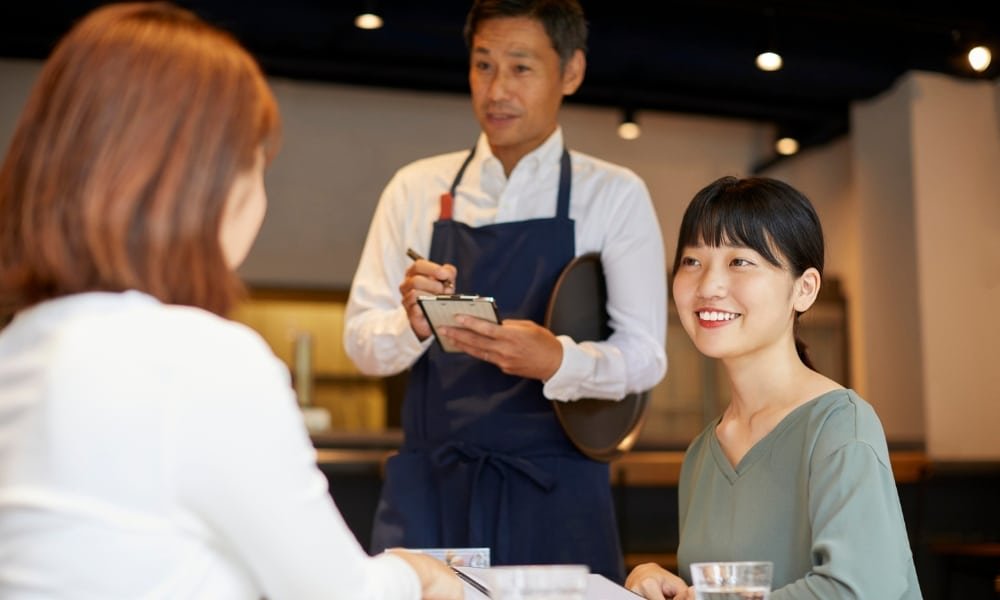 พนักงานเสิร์ฟในผ้ากันเปื้อนรับออเดอร์จากผู้หญิงสองคนที่นั่งอยู่ที่ ที่เที่ยวกรุงเทพ โต๊ะในร้านอาหาร ซึ่งเป็นหนึ่งในสถานที่ท่องเที่ยวยอดนิยมในกรุงเทพฯ สำหรับการรับประทานอาหารและพักผ่อน.