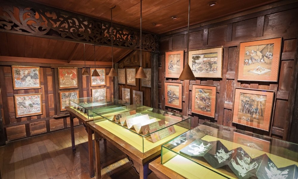 ห้องพิพิธภัณฑ์ที่มีผนังไม้จัดแสดงผลงานศิลปะและวัตถุโบราณบนกระดาษในตู้กระจกใต้แสงไฟระยิบ ที่เที่ยวกรุงเทพ ระยับ—สถานที่ท่องเที่ยวในกรุงเทพฯ ที่สร้างแรงบันดาลใจสำหรับผู้รักประวัติศาสตร์และศิลปะ.