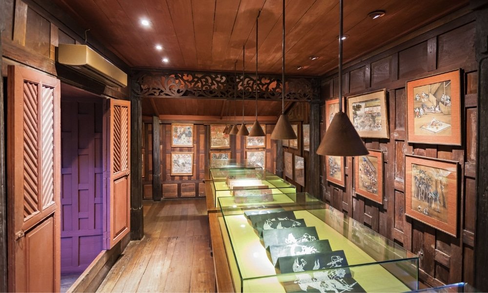 แกลเลอรีไม้ที่มีแผงไม้ล้อมรอบ  ที่เที่ยวกรุงเทพ พร้อมผลงานศิลปะที่ติดกรอบและตู้โชว์ ซึ่งส่องสว่างด้วยโคมไฟแขวน—เหมาะสำหรับผู้ที่ต้องการสัมผัสประสบการณ์ท่องเที่ยวกรุงเทพฯ ที่ไม่เหมือนใคร.