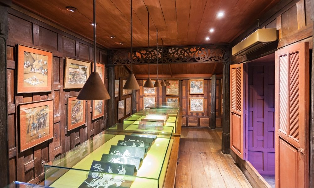 ห้องพิพิธภัณฑ์ที่ตกแต่งด้วยไม้หนึ่งในสถานที่ท่องเที่ยวกรุงเทพมหานครชั้น ที่เที่ยวกรุงเทพ ยอด แสดงผลงานศิลปะในกรอบบนผนังและหุ่นเงาในตู้กระจกใต้แสงไฟที่แขวนอยู่.