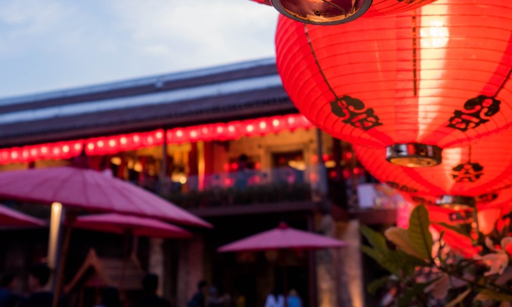โคมไฟจีนสีแดงแขวนอยู่กลางแจ้งพร้อมฉากหลังของตึกที่เบลอและโคมไฟเพิ่มเติม สร้างบรรยากาศที่ครึกครื้น—เหมาะสำหรับผู้ที่ต้องการสถานที่ท่องเที่ยวในกรุงเทพฯ ที่ถ่ายรูปสวยๆ ใน ที่เที่ยวกรุงเทพ กรุงเทพฯ.