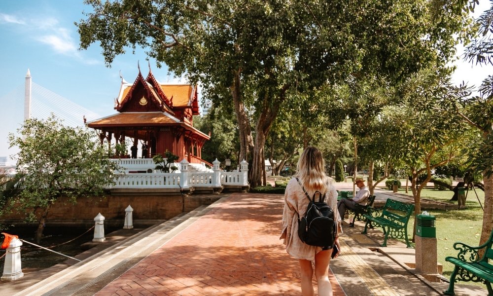 ผู้หญิงคนหนึ่งสะพายกระเป๋าเป้เดินไปตามทางเดินอิฐมุ่งหน้าไปยัง ที่เที่ยวกรุงเทพ ศาลาไทยแบบดั้งเดิมที่ล้อมรอบด้วยต้นไม้และม้านั่งในสวนสาธารณะ กำลังสำรวจหนึ่งในสถานที่ท่องเที่ยวที่เงียบสงบในกรุงเทพฯ.