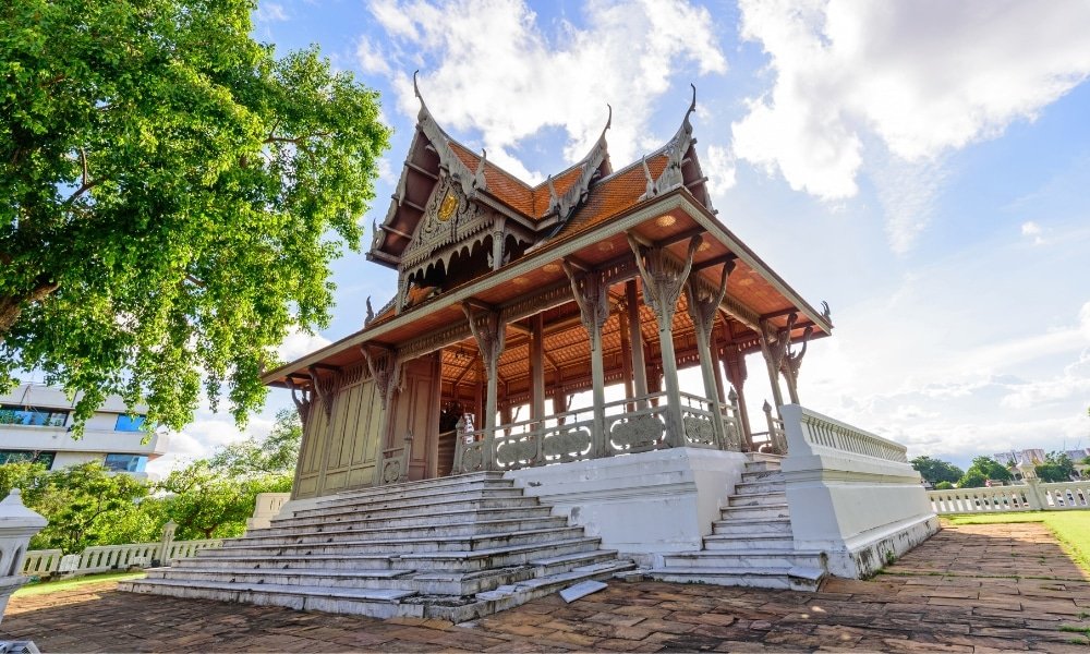 ศาลาไทยแบบดั้งเดิมพร้อมหลังคาไม้แกะสลักอย่างวิจิตรและลวดลายประดับประดา ตั้งอยู่บนบันไดกว้างสีขาว รายล้อมด้วยต้นไม้สีเขียวภาย ที่เที่ยวกรุงเทพ ใต้ท้องฟ้าที่มีเมฆบางส่วน—สถานที่ท่องเที่ยวในกรุงเทพฯ ที่น่าดึงดูดใจสำหรับนักท่องเที่ยวที่แสวงหาเสน่ห์ทางวัฒนธรรม.