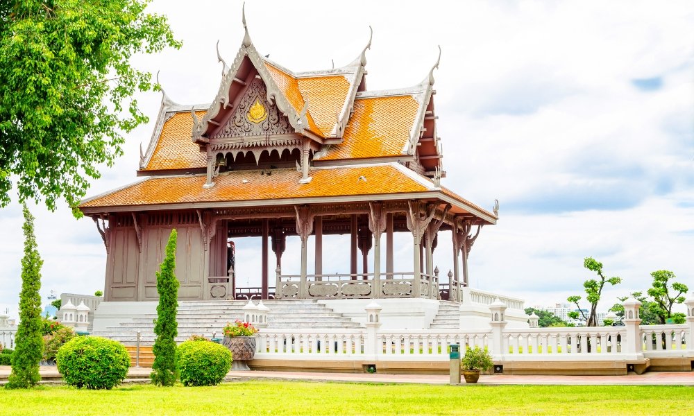 ศาลาไทยแบบดั้งเดิมหลังคาประดับประดาอย่างวิจิตร พร้อม ที่เที่ยวกรุงเทพ กระเบื้องสีส้ม ล้อมรอบด้วยต้นไม้สีเขียวและรั้วสีขาว—สถานที่ท่องเที่ยวกรุงเทพที่เป็นสัญลักษณ์ ท่ามกลางท้องฟ้าที่มีเมฆปกคลุม.