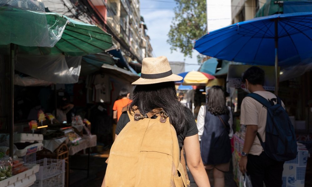 บุคคลที่สวมหมวกและเป้สะพาย ที่เที่ยวกรุงเทพ หลังกำลังสำรวจตลาดกลางแจ้งที่คึกคัก หนึ่งในสถานที่ท่องเที่ยวที่เต็มไปด้วยชีวิตชีวาในกรุงเทพฯ รายล้อมไปด้วยแผงขายของและผู้คนที่อยู่ใต้ร่มหลากสีสัน.