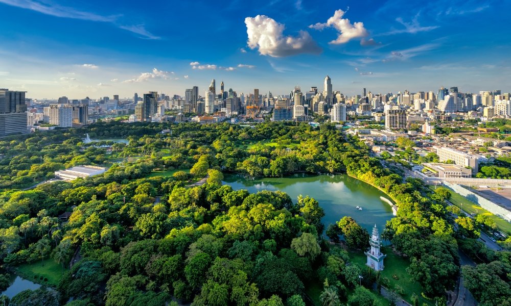 ภาพมุมสูงของสวนสาธารณะขนาดใหญ่สีเขียว มีทะเลสาบ เป็นหนึ่งในสถานที่ท่องเที่ยวที่ดีที่สุดในกรุงเทพฯ ล้อมรอบด้วยตึกสูงใน ที่เที่ยวในกรุงเทพ เมืองภายใต้ท้องฟ้าสีครามที่มีเมฆกระจายอยู่.