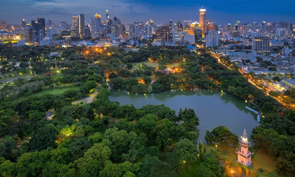 ภาพมุมสูงของสวนสาธารณะขนาดใหญ่ในเมืองใหญ่ มีทะเลสาบ ล้อมรอบด้วยต้นไม้และทางเดินที่ ที่เที่ยวในกรุงเทพ สว่างไสว พร้อมทัศนียภาพของกรุงเทพมหานครที่สวยงามตระการตาในยามพลบค่ำเป็นฉากหลัง.