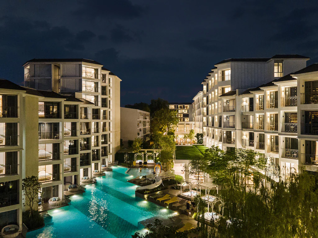 ภาพยามค่ำคืนของรีสอร์ทสมัยใหม่ในแหล่งท่องเที่ยวกรุงเทพมหานคร พร้อมอาคารที่สว่างไสวและสระว่ายน้ำกลางแจ้งขนาดใหญ่ ล้อมรอบด้วยเก้าอี้เลานจ์และพืชพรรณเขียว ที่เที่ยวกรุงเทพ ชอุ่ม.