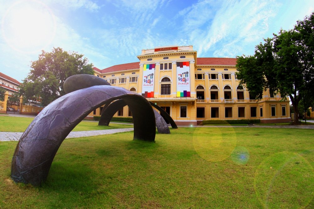 อาคารสีเหลืองขนาดใหญ่พร้อมป้ายแบนเนอร์และหน้าต่างโค้งตั้งอยู่ข้างประติมากรรมสีดำสมัยใหม่บนสนามหญ้าใต้ท้องฟ้าที่มีเมฆบางส่วน—สถานที่ท่องเที่ยวอันเป็นสัญลักษณ์ของกรุงเทพมหานคร ที่เที่ยวกรุงเทพ สำหรับผู้ที่กำลังสำรวจอัญมณีทางสถาปัตยกรรมของเมือง.