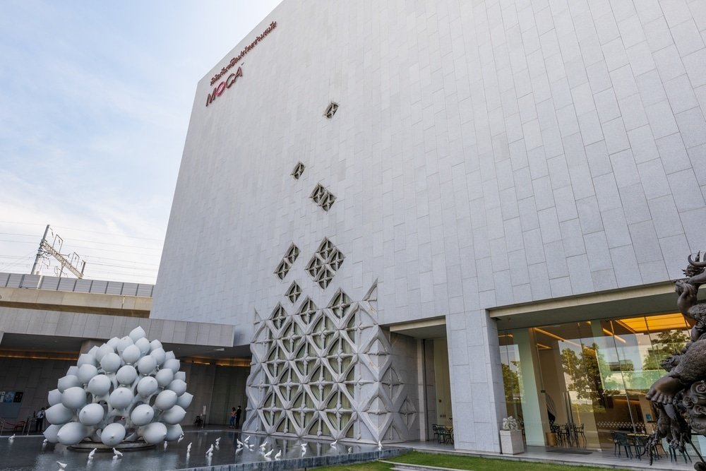 อาคารสีขาวสมัยใหม่ที่มีลวดลายผนังเรขาคณิต ประติมากรรมทรงกลมสีขาวกลุ่มหนึ่ง และสระน้ำ ที่เที่ยวกรุงเทพ สะท้อนแสงอยู่ด้านหน้า ป้ายเขียนว่า "MOCA"—สถานที่ท่องเที่ยวในกรุงเทพฯ ที่ต้องไปเยือนสำหรับคนรักศิลปะที่กำลังสำรวจเมือง.