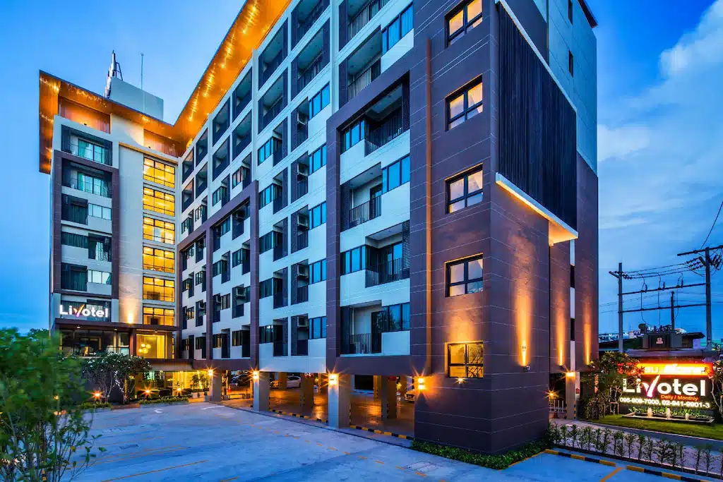 อาคารโรงแรมหลายชั้นสมัยใหม่ โรงแรมในกรุงเทพ พร้อมไฟส่องสว่างภายนอก หน้าต่างขนาดใหญ่ และพื้นที่จอดรถด้านหน้า ป้ายแสดงชื่อโรงแรม "Livotel" มองเห็นได้ชัดเจน—ที่พักในกรุงเทพฯ ที่เหมาะสำหรับความสะดวกสบายและความสะดวกในการเดินทาง.