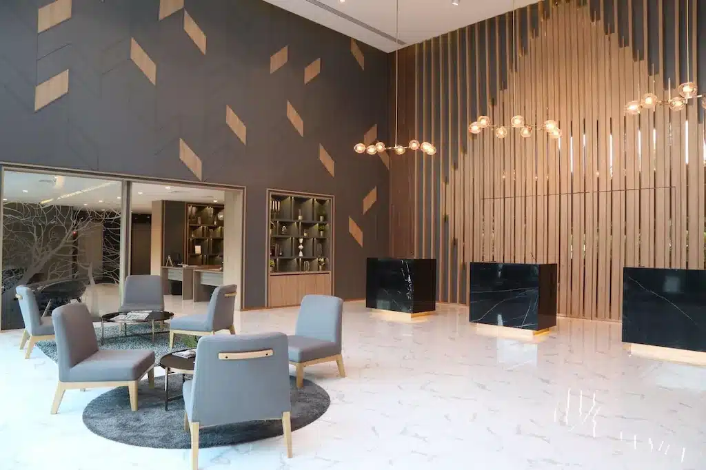 ล็อบบี้โรงแรมสมัยใหม่ในโรงแรม 5 ดาวกรุงเทพฯ ประกอบด้วยเก้าอี้สี โรงแรมในกรุงเทพ เทา โต๊ะเล็ก พื้นหินอ่อน ลวดลายผนังเรขาคณิต และเคาน์เตอร์ต้อนรับสีดำสามตัวที่มีแผ่นไม้และโคมไฟแขวนอยู่ด้านหลัง.