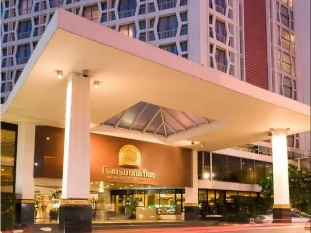 ทางเข้าของอาคารโรงแรมสมัยใหม่ โรงแรมในกรุงเทพ ฯ มีเสาขนาดใหญ่ ประตูกระจก และป้ายเหนือทางเข้าที่เขียนเป็นภาษาไทยและภาษาอังกฤษ ต้อนรับแขกสู่โรงแรมกรุงเทพที่มีสไตล์แห่งนี้.