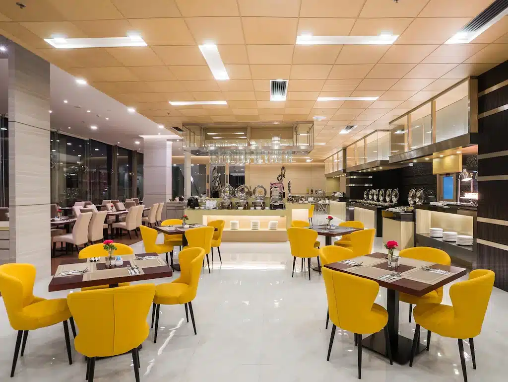 การตกแต่งภายในร้านอาหารสมัยใหม่ที่ โรงแรม 5 ดาวในกรุงเทพ ฯ พร้อมเก้าอี้สีเหลือง โต๊ะที่จัดเรียงอย่างเรียบร้อย เคาน์เตอร์บุฟเฟ่ต์ และแสงไฟเหนือศีรษะที่สว่างสดใส.