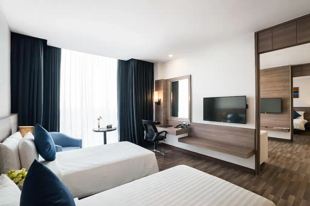ห้องพักโรงแรมสมัยใหม่ที่ โรงแรมกรุงเทพ พร้อมเตียงเดี่ยวสองเตียง โต๊ะและเก้าอี้ ทีวีจอแบน หน้าต่างบานใหญ่พร้อมผ้าม่าน และโต๊ะกลมขนาดเล็กพร้อมเก้าอี้.
