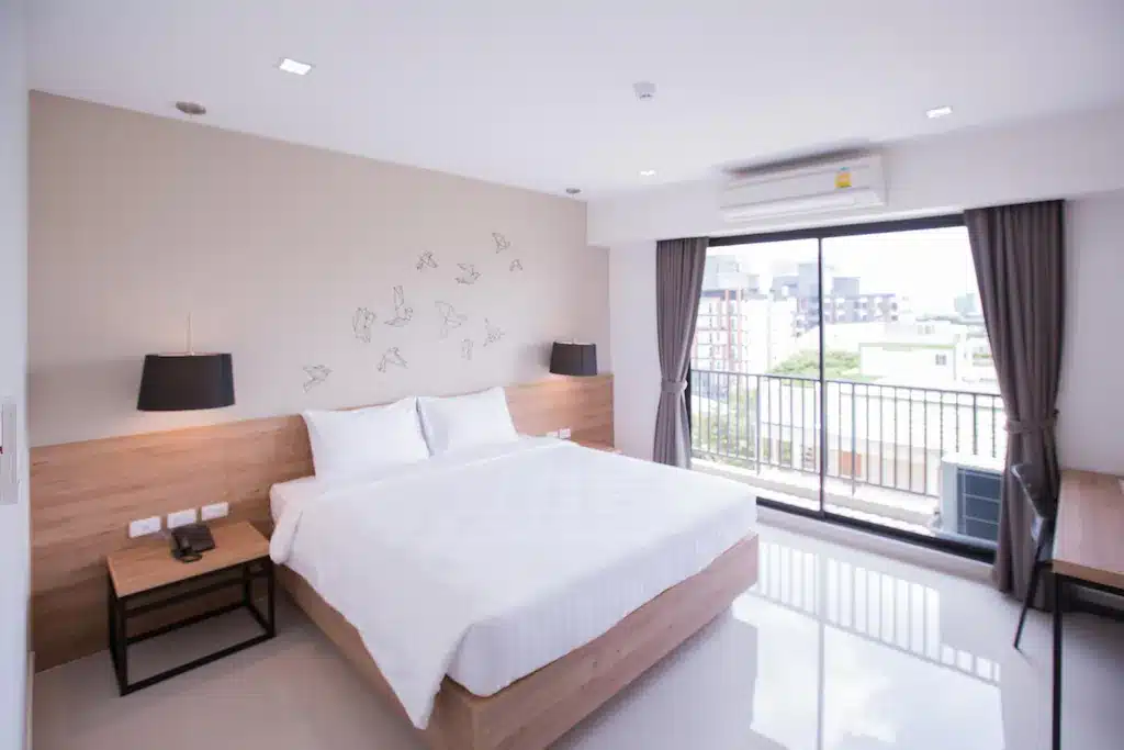 ห้องพักโรงแรมสมัยใหม่พร้อมเตียงขนาดใหญ่ โต๊ะข้างเตียง ศิลปะบนผนัง โต๊ะทำงาน และหน้าต่างบาน โรงแรมในกรุงเทพ ใหญ่จากพื้นจรดเพดานพร้อมวิวระเบียง พื้นที่ที่มีสไตล์นี้สะท้อนถึงความหรูหราของโรงแรม 5 ดาวในกรุงเทพฯ พร้อมการตกแต่งที่สว่างและโทนสีเรียบง่าย.