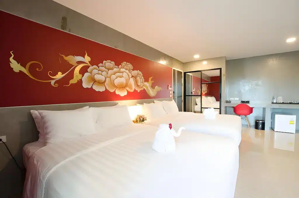 ห้องพักโรงแรมสมัยใหม่ในโรงแรม 5 ดาวกรุงเทพ พร้อมเตียงใหญ่สอง โรงแรมกรุงเทพ เตียง ผนังสีแดงตัดกับศิลปะดอกไม้ ผ้าปูที่นอนสีขาว ผ้าเช็ดตัวพับเป็นรูปสัตว์ โต๊ะทำงานพร้อมเก้าอี้สีแดง และตู้เย็นขนาดเล็ก.