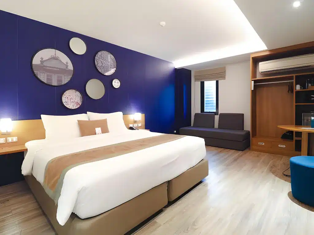 ห้องพักโรงแรมสมัยใหม่ที่ โรงแรม 5 ดาวกรุงเทพ ประกอบด้วยเตียงขนาดใหญ่ ผนังสีน้ำเงินเป็นจุดเด่น งาน โรงแรมกรุงเทพ ศิลปะทรงกลม โคมไฟติดผนัง โซฟา เครื่องปรับอากาศ พื้นไม้ และโต๊ะทำงานและที่เก็บของแบบบิลท์อิน.