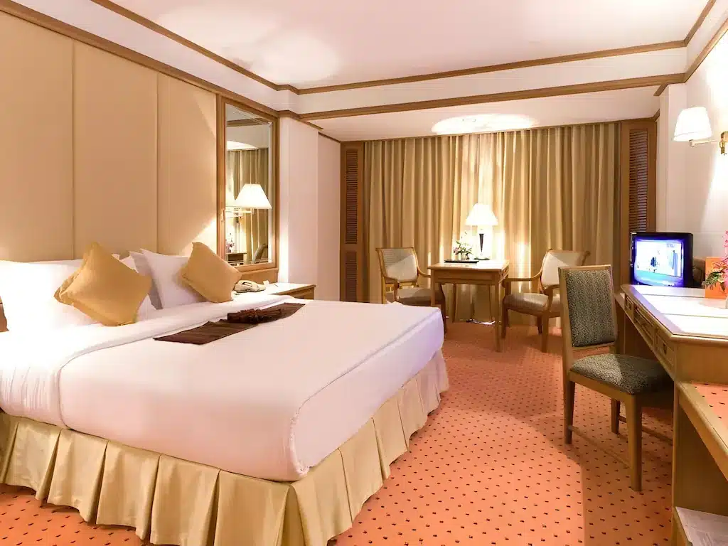 ที่พักกรุงเทพสไตล์โมเดิร์น พร้อม โรงแรมในกรุงเทพ เตียงขนาดใหญ่, โต๊ะข้างเตียงสองตัวพร้อมโคมไฟ, โต๊ะทำงานและเก้าอี้, โทรทัศน์ขนาดเล็กบนตู้ลิ้นชัก, และมุมนั่งเล่นสบาย ๆ ใกล้หน้าต่างพร้อมผ้าม่าน—เหมาะสำหรับการพักผ่อนในทริปต่อไปของคุณ.