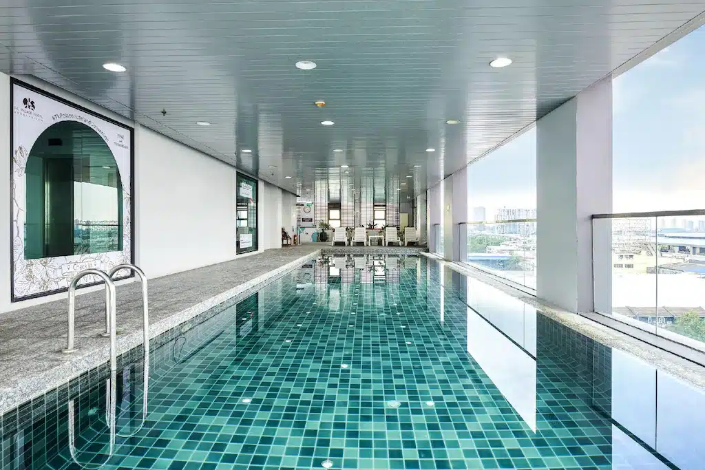 สระว่ายน้ำในร่มพร้อมพื้นกระเบื้อง บันไดสแตนเลส และหน้าต่างบานใหญ่ที่มองเห็นวิวเมือง ณ โรงแรม 5 ดาว กรุงเทพ มีโต๊ะและเก้าอี้ตั้งอยู่ปลายสุดของที่พักหรูแห่งนี้.