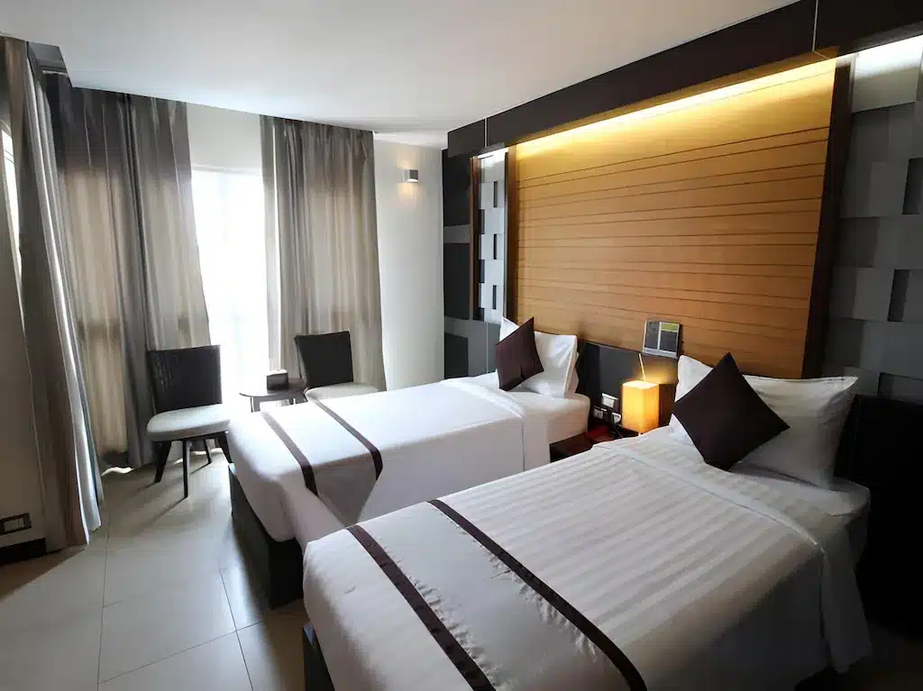 ห้องพักโรงแรมสมัยใหม่ที่ โรงแรมกรุงเทพ พร้อมเตียงเดี่ยวสองเตียง ผ้าปูที่นอนสีขาว หมอนตกแต่งสีเข้ม หัวเตียงไม้สัก โต๊ะเล็ก เก้าอี้สองตัว และหน้าต่างบานใหญ่พร้อมผ้าม่าน.