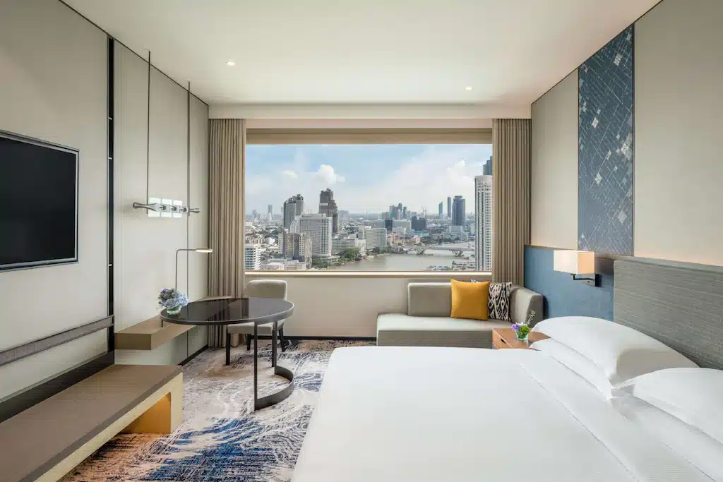 ห้องพักโรงแรมสมัยใหม่พร้อมเตียงขนาดใหญ่ โซฟา โต๊ะทำงาน และทีวีจอแบน; หน้าต่างบานใหญ่ให้วิวเส้นขอบฟ้าเมืองและแม่น้ำ—เหมาะ โรงแรมกรุงเทพ สำหรับผู้ที่ต้องการที่พักในกรุงเทพฯ ในโรงแรม 5 ดาวกรุงเทพฯ ในเวลากลางวัน.