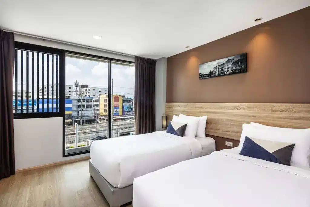 โรงแรมในกรุงเทพ ห้องพร้อมเตียงคู่สองเตียง ผนังสีน้ำตาลเป็นจุดเด่น หน้าต่างบานใหญ่พร้อมวิวเมือง พื้นไม้ และภาพถ่ายขาวดำเหนือหัวเตียง.
