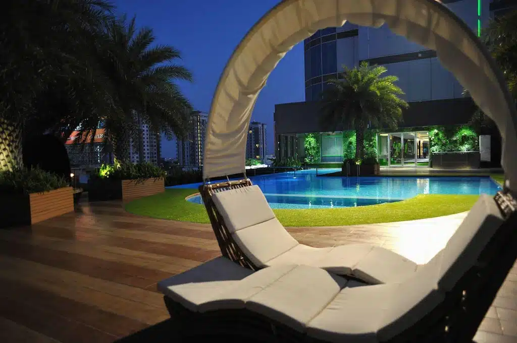 พื้นที่สระว่ายน้ำกลางแจ้งสมัยใหม่ยามเย็นที่ โรงแรมกรุงเทพ พร้อมเก้าอี้เลานจ์โค้งใต้หลังคาสีขาว ดาดฟ้าไม้ ต้น โรงแรมในกรุงเทพ ปาล์ม และตึกสูงในเมืองเป็นฉากหลัง.