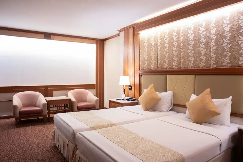 โรงแรม 5 ดาวกรุงเทพ ห้องพักมี โรงแรมในกรุงเทพ เตียงคู่ ตกแต่งโทนเบจทอง พร้อมโต๊ะหัวเตียงและโคมไฟ มุมที่นั่งเล็กๆ กับเก้าอี้สองตัวและโต๊ะริมหน้าต่าง เหมาะสำหรับที่พักกรุงเทพสุดหรูใจกลางเมือง.