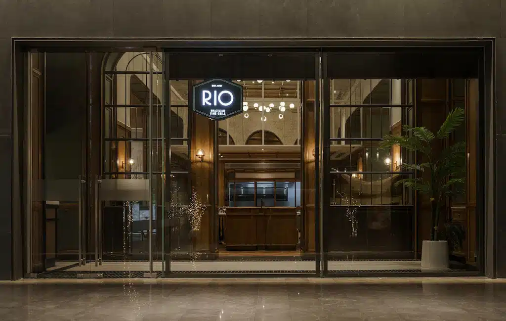 ทางเข้าโรงแรมในกรุงเทพฯ ที่ทำจากกระจก มีป้ายไฟ "RIO" ส่อง โรงแรมในกรุงเทพ สว่างอยู่เหนือเคาน์เตอร์ต้อนรับไม้ ภายในมีต้นไม้ในกระถางและไฟสมัยใหม่ มองเห็นได้อย่างชัดเจน มอบการต้อนรับอย่างมีสไตล์สำหรับการเข้าพักในเมืองของคุณ.