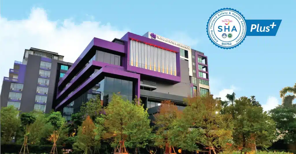 อาคารหลายชั้นสมัยใหม่พร้อมการตกแต่งสีม่วงล้อมรอบด้วยต้นไม้ ท่ามกลางท้องฟ้าสีคราม เหมาะสำหรับผู้ที่มองหาโรงแรมกรุงเทพหรือที่พักหรูหราในกรุงเทพมหานคร. แสดงตราสัญลักษณ์ "SHA Plus+" ที่มุมขวาบน.