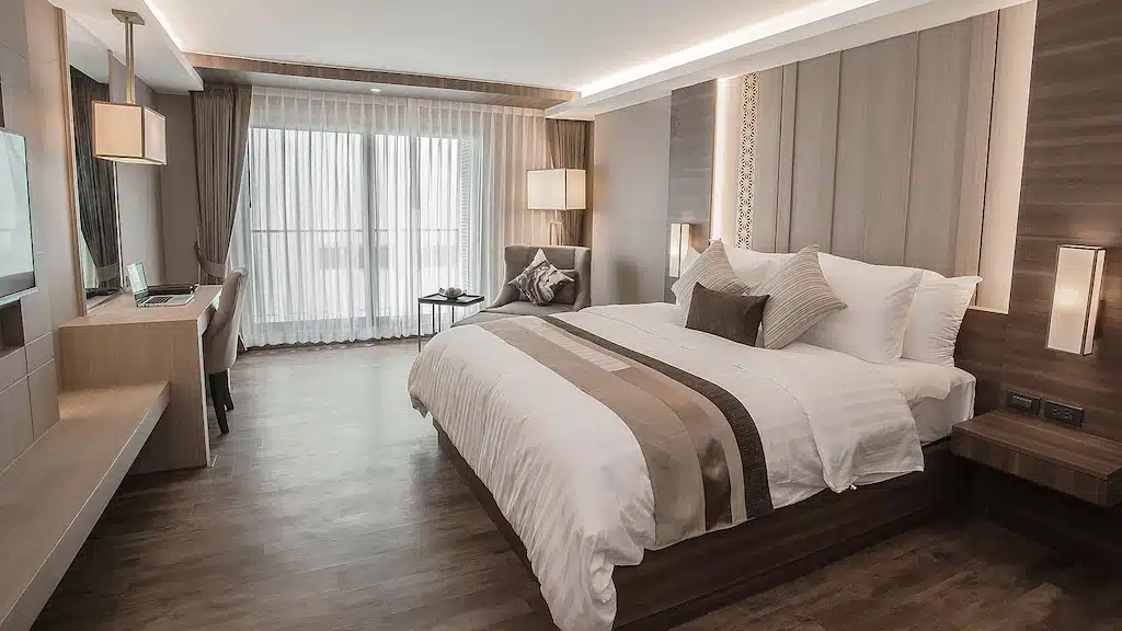 ห้องพักโรงแรมสมัยใหม่ใน โรงแรม 5 ดาวกรุงเทพ , พร้อมเตียงขนาดใหญ่, ตกแต่งสไตล์กลาง, โต๊ะทำงานและเก้าอี้, โต๊ะเล็กสำหรับนั่งเล่น, พื้นไม้, และหน้าต่างขนาดใหญ่พร้อมผ้าม่านบาง.