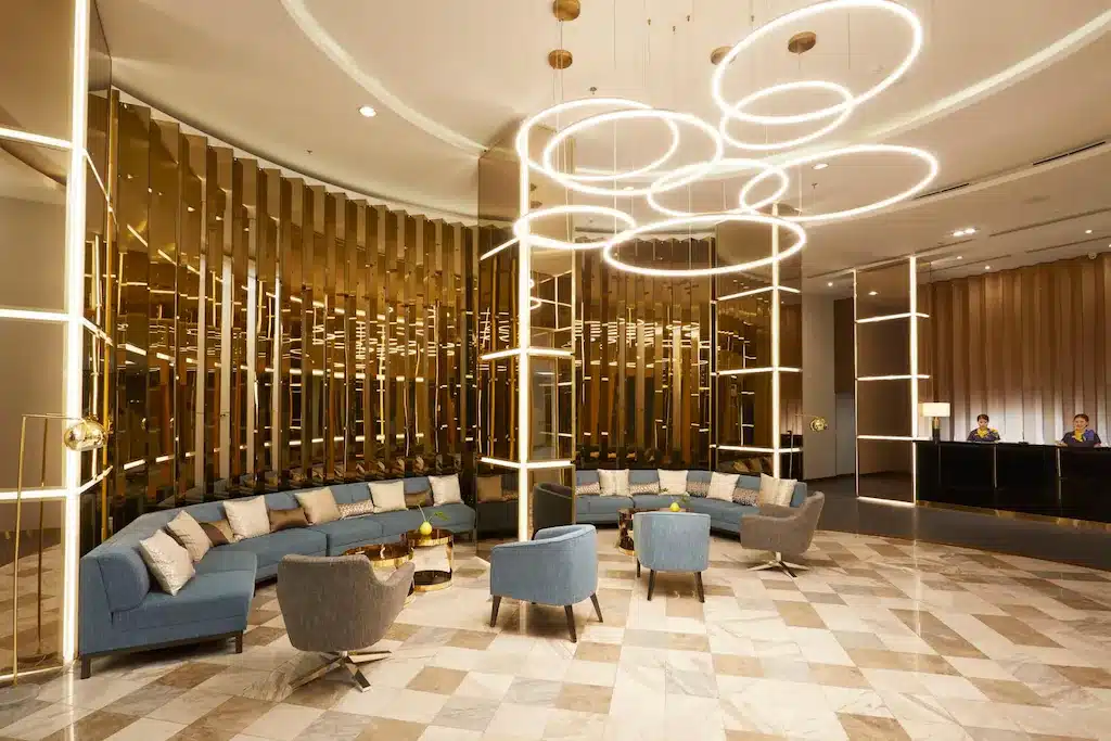 ล็อบบี้โรงแรมสมัยใหม่ในโรงแรม 5 ดาวกรุงเทพ, โดดเด่นด้วยที่นั่ง โรงแรมกรุงเทพ สีน้ำเงินและเทา, พื้นหินอ่อน, รายละเอียดสีทอง, และโคมไฟแขวนทรงกลม. โต๊ะต้อนรับพร้อมพนักงานสองคนสามารถมองเห็นได้ในพื้นหลัง.