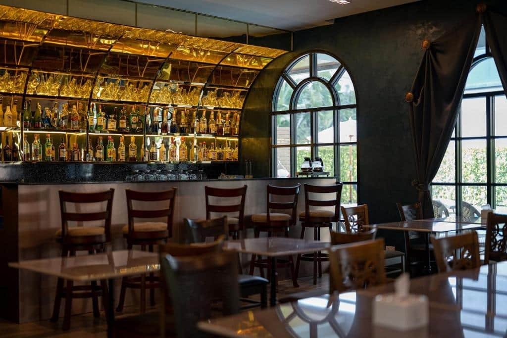 บาร์สมัยใหม่ที่โรงแรม 5 ดาวในกรุงเทพฯ มีเก้าอี้พนักสูง ผนังด้านหลังเป็นกระจกเงาและไฟสีทองประดับขวดเหล้าเรียงราย และหน้าต่างโค้งขนาดใหญ่พร้อมผ้าม่านที่ห้อยลงมา.