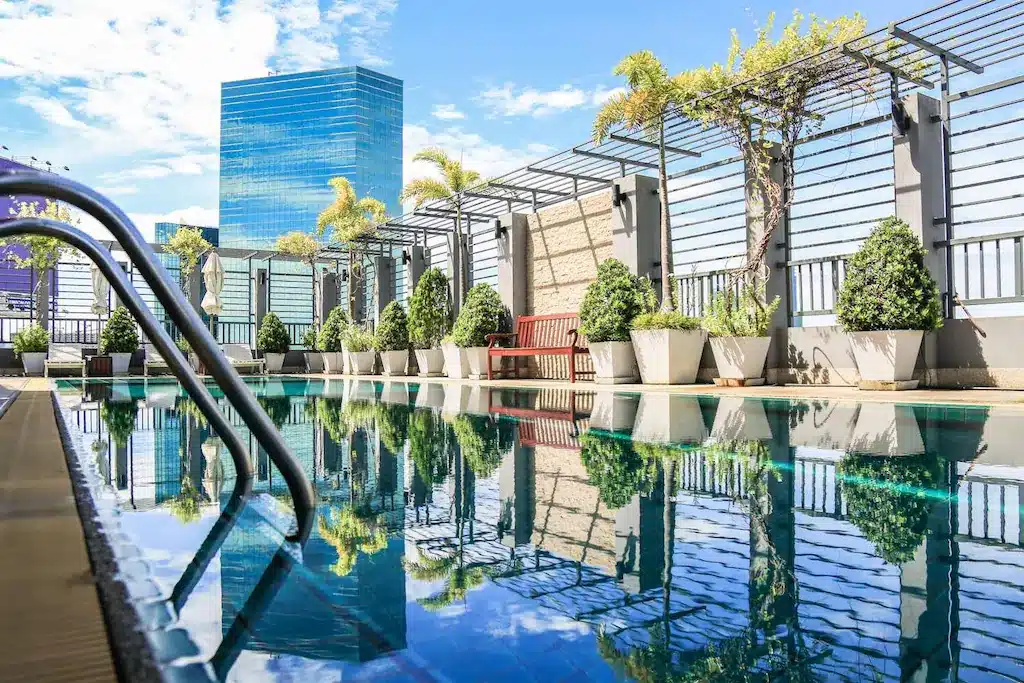 สระว่ายน้ำบนดาดฟ้าของ โรงแรมในกรุงเทพ ฯ ล้อมรอบด้วยต้นไม้ในกระถางและม้านั่งไม้ พร้อมวิวตึกระฟ้าของเมืองที่มองเห็นอยู่เบื้องหลังภายใต้ท้องฟ้าสีฟ้าใส.