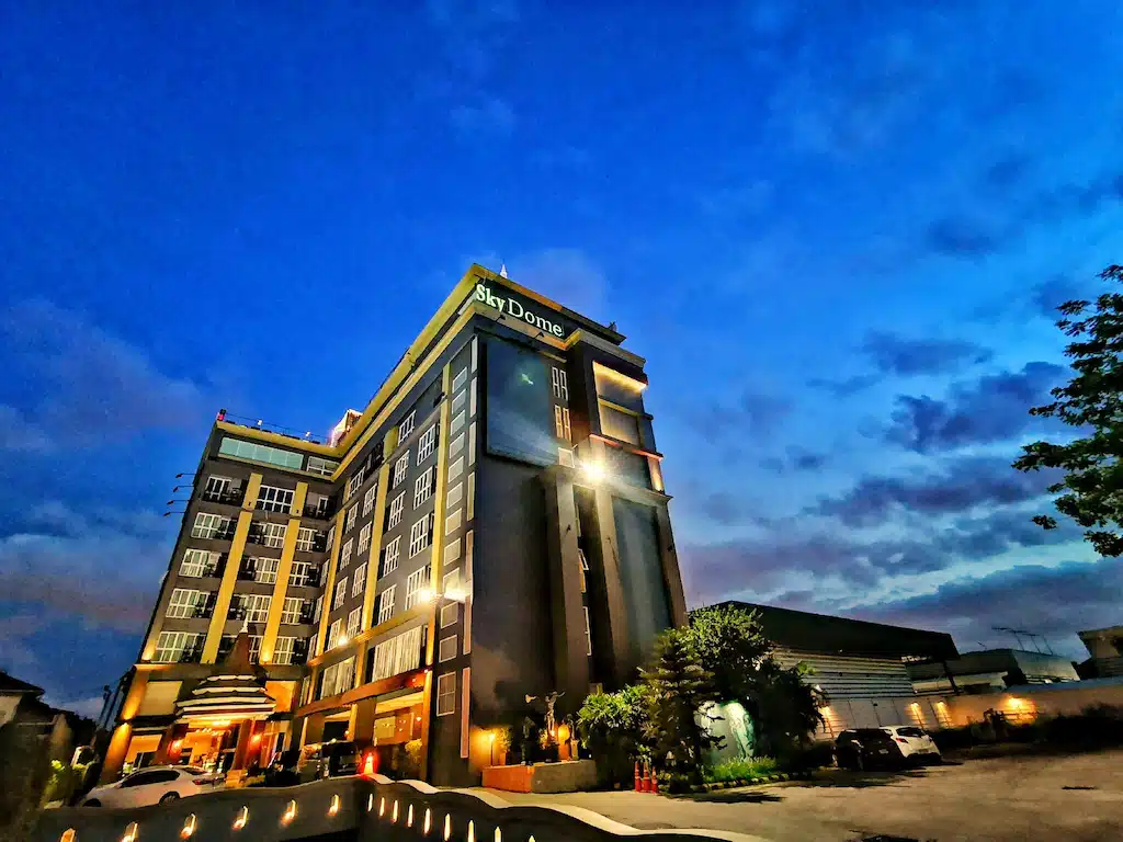 อาคารหลายชั้นที่มีป้ายชื่อว่า  โรงแรมกรุงเทพ "SkyDome" ถูกส่องสว่างในยามพลบค่ำ ท้องฟ้าแจ่มใสและมีเมฆกระจายอยู่เป็นระยะๆ รถยนต์จอดอยู่หน้าโรงแรมระดับ 5 ดาวในกรุงเทพฯ แห่งนี้ ซึ่งมอบการพักผ่อนที่หรูหราใจกลางเมือง.