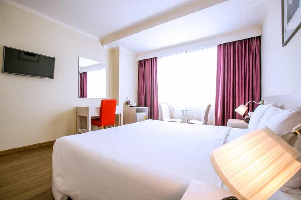 ห้องพักโรงแรมในกรุงเทพ ฯ ที่สว่างสดใสพร้อมเตียงคู่ ผ้าปูที่นอนสีขาวสะอาด โต๊ะทำงานพร้อมเก้าอี้สีส้ม โทรทัศน์ติดผนัง และหน้าต่างบานใหญ่พร้อมผ้าม่านสีแดง—เหมาะสำหรับการเข้าพักครั้งต่อไปของคุณในเมือง.