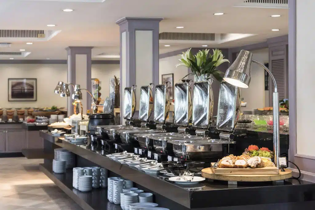 การจัดบุฟเฟ่ต์ของโรงแรมในกรุงเทพฯ มีถาดอุ่นอาหาร, อุปกรณ์เสิร์ฟ, จานซ้อน, และอาหารหลากหลายชนิดที่จัดเรียงอย่างเรียบร้อยบนเคาน์เตอร์ยาว. โรงแรมกรุงเทพ 