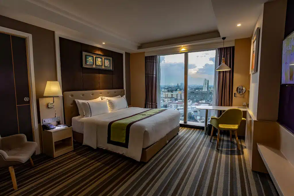 ห้องพักโรงแรมสมัยใหม่พร้อมเตียงขนาดใหญ่ พื้นที่โต๊ะทำงาน และ โรงแรมในกรุงเทพ หน้าต่างบานใหญ่จากพื้นจรดเพดานที่มองเห็นทิวทัศน์เมืองยามพระอาทิตย์ตก แสงไฟอบอุ่นและการตกแต่งร่วมสมัยสร้างบรรยากาศที่แสนสบาย—เหมาะสำหรับการเข้าพักของคุณที่ โรงแรมกรุงเทพ.