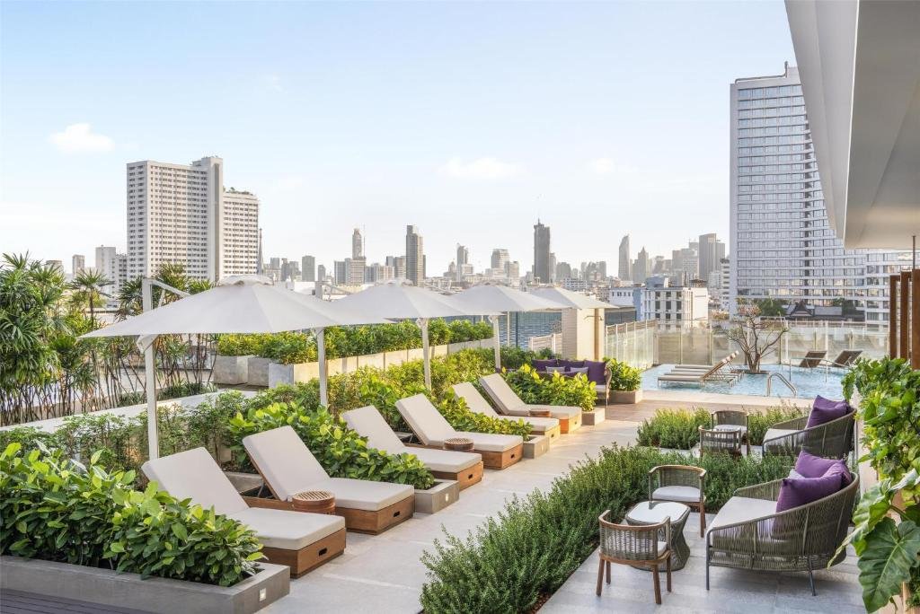 สระว่ายน้ำบนดาดฟ้าของโรงแรม 5 ดาวในกรุงเทพฯ พร้อมเก้าอี้อาบแดด ร่มไม้ร่มรื่น และวิวเส้นขอบฟ้าเมืองที่สวยงามในวันที่อากาศแจ่มใส. โรงแรมกรุงเทพ 