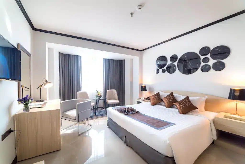 ห้องพักโรงแรมสมัยใหม่ที่ โรงแรม 5 ดาวในกรุงเทพ ฯ พร้อมเตียงขนาดใหญ่ เก้าอี้สองตัวข้างหน้าต่าง โต๊ะทำงานพร้อมโคมไฟ และงานศิลปะผนังวงกลมสมัยใหม่เหนือหัวเตียง.
