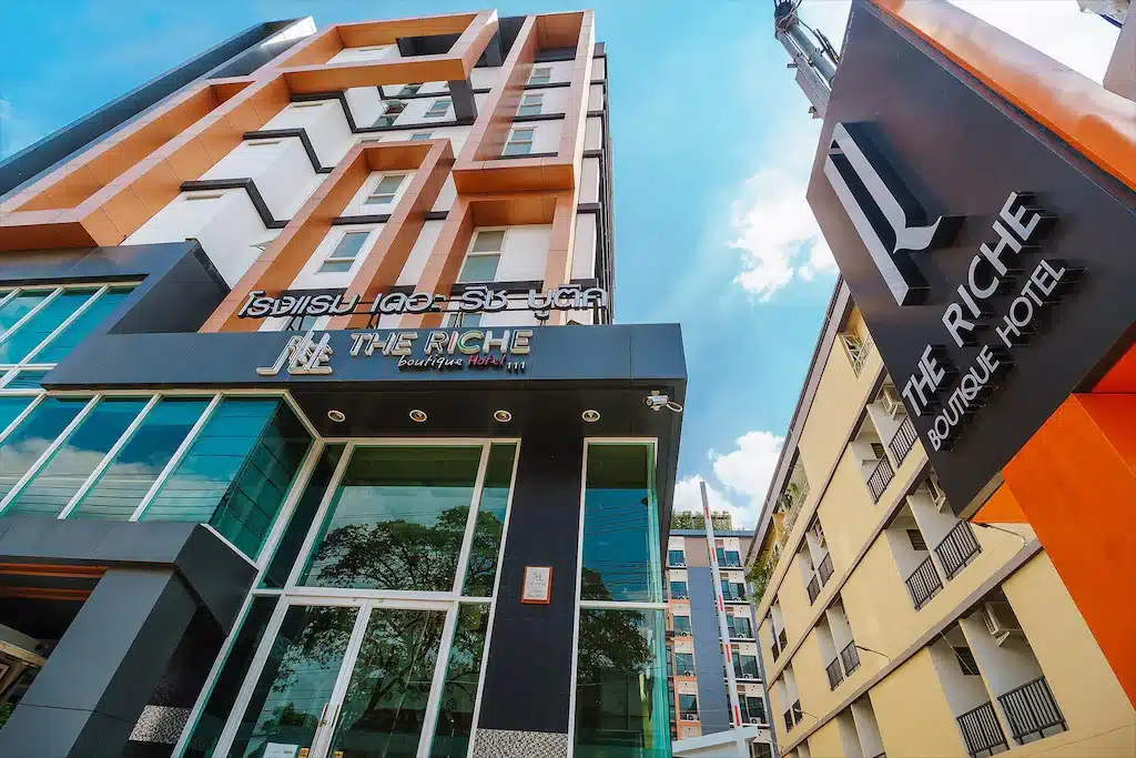 อาคารหลายชั้นสมัยใหม่พร้อมป้าย "The Riche Boutique Hotel" ทาง โรงแรมกรุงเทพ เข้าเป็นกระจก และมีป้ายบอกทางอยู่ติดกันภายใต้ท้องฟ้าสีฟ้า—ทางเลือกที่น่าดึงดูดสำหรับผู้ที่ต้องการ โรงแรมกรุงเทพ หรือ ที่พักกรุงเทพ.