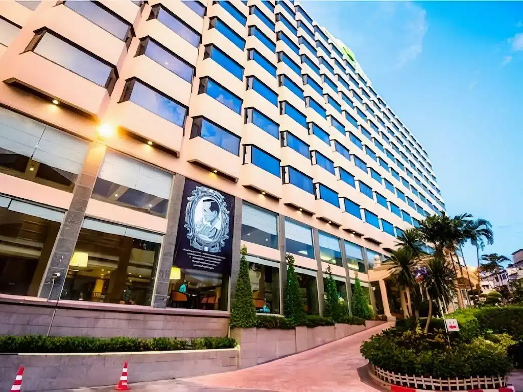 โรงแรม 5 ดาวในกรุงเทพ ฯ ทันสมัยหลายชั้น พร้อมหน้าต่างบานใหญ่ ภูมิทัศน์สีเขียว และทางเข้าด้านหน้า ถ่ายจากมุมต่ำภายใต้ท้องฟ้าแจ่มใส.
